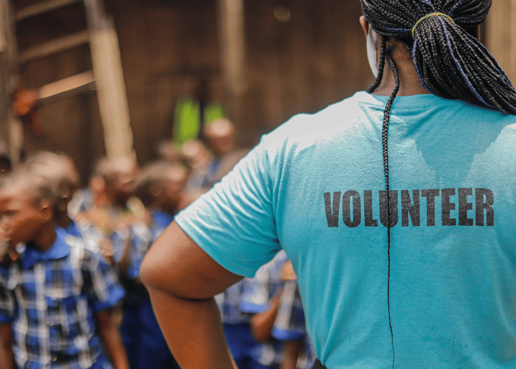 A volunteer in a teal shirt stands with her back to the camera, facing a group of children in school uniforms. The word "Volunteer" is printed across the back of her shirt, and the children are softly blurred in the background.