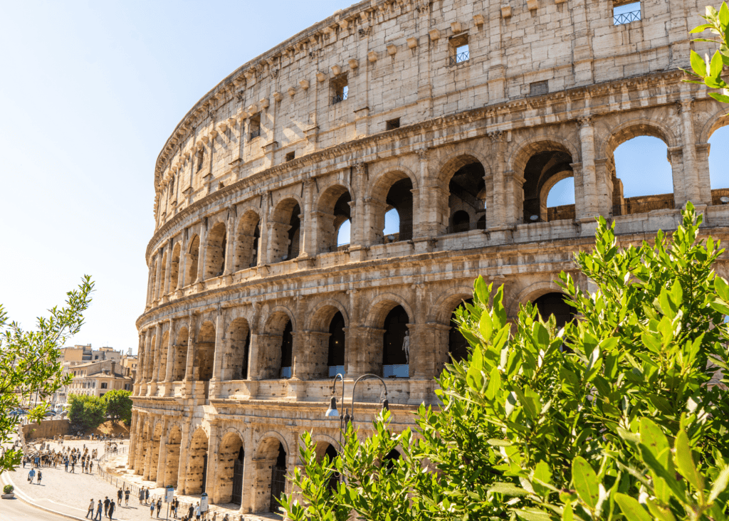 The ancient exterior facade of the Roman Colosseum rises against a clear blue sky in Rome, Italy, its massive arched stonework framed by green trees with visitors gathered along the street below.