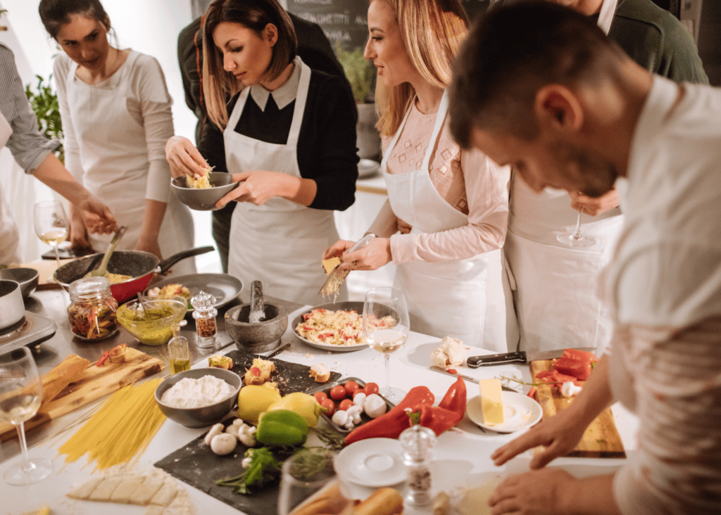 A group of travelers in white aprons gather around a kitchen counter during an Italian cooking class, preparing fresh pasta and vegetables alongside wine in a warmly lit culinary studio in Florence.