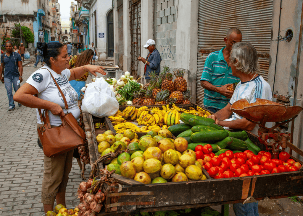 A woman shops at a vibrant outdoor street market in Colombia, selecting fresh produce including bananas, pineapples, tomatoes, and cucumbers from a wooden cart vendor on a cobblestone street.