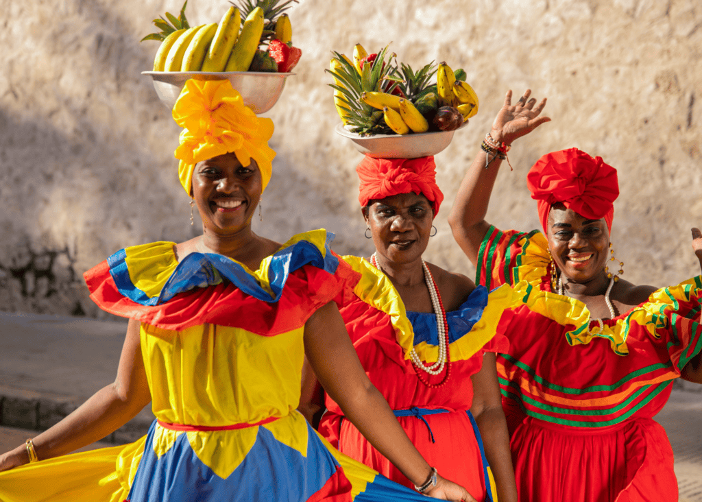 Three joyful Palenquera women in vibrant ruffled dresses in the colors of the Colombian flag balance bowls of fresh tropical fruit on their heads against the ancient stone walls of Cartagena's old city.