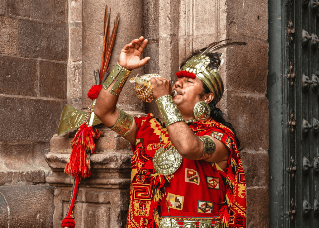 A man dressed in elaborate traditional Inca ceremonial clothing including a gold headdress with feathers, ornate bronze cuffs, and a vibrant red woven tunic plays a conch shell outside a stone cathedral in Cusco, Peru.