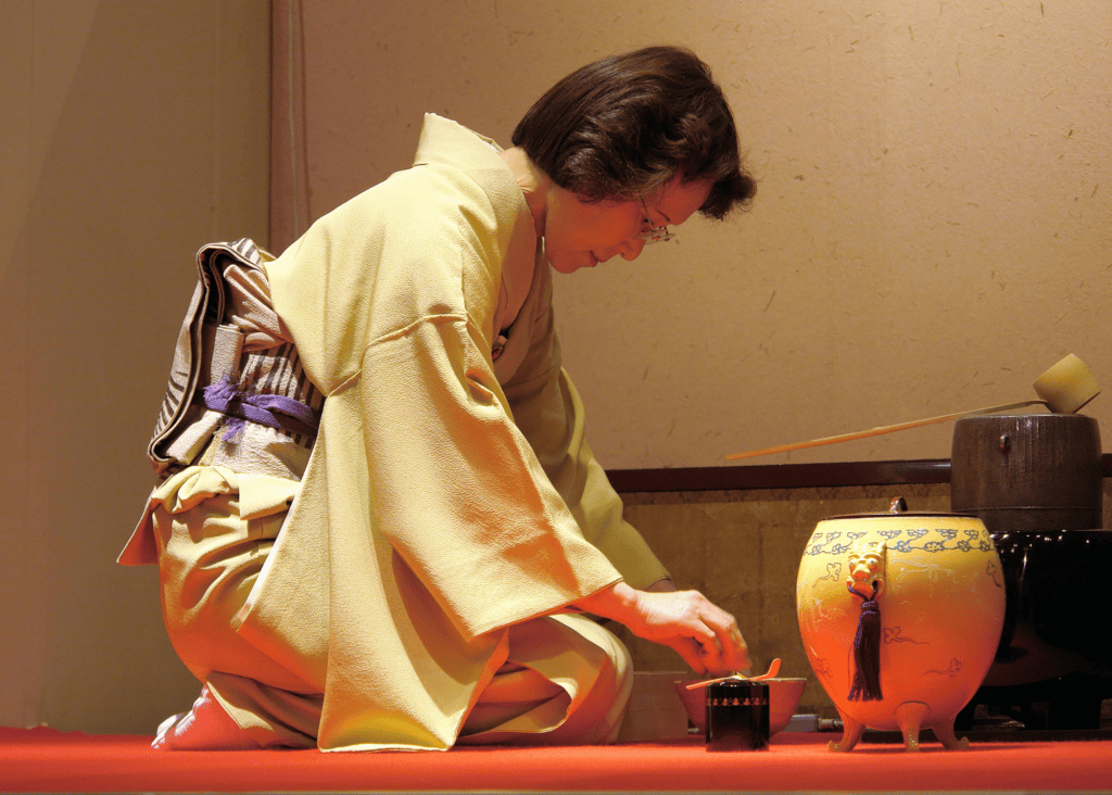 A woman dressed in a pale yellow kimono kneels gracefully while performing a traditional Japanese tea ceremony, carefully preparing matcha with handcrafted ceramic vessels and lacquered tools.