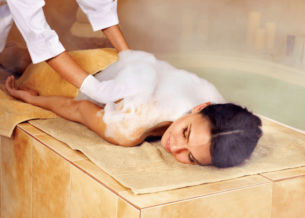A woman relaxes peacefully on a warm marble slab while a therapist performs a traditional Turkish hammam scrub treatment, covering her back in rich lather in a steam filled bath house.