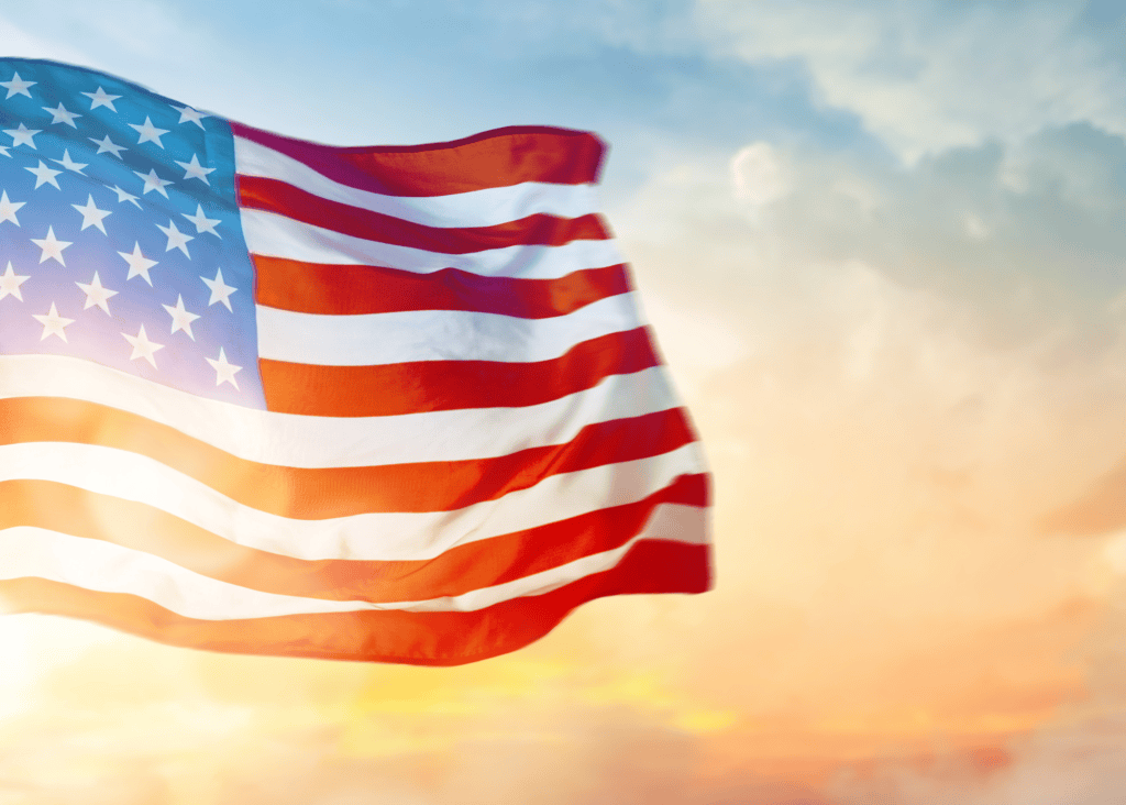 The American flag waves proudly against a glowing orange and gold sunset sky, its red white and blue colors illuminated in the warm light of dusk.