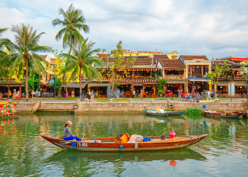 A woman rows a traditional wooden boat along the Thu Bon River in Hoi An, Vietnam, with the colorful lantern-lit buildings and palm trees of the ancient town reflecting in the calm water behind her.