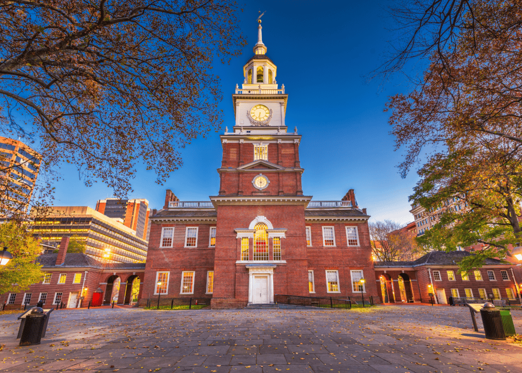 The American flag waves proudly against a glowing orange and gold sunset sky, its red white and blue colors illuminated in the warm light of dusk.
