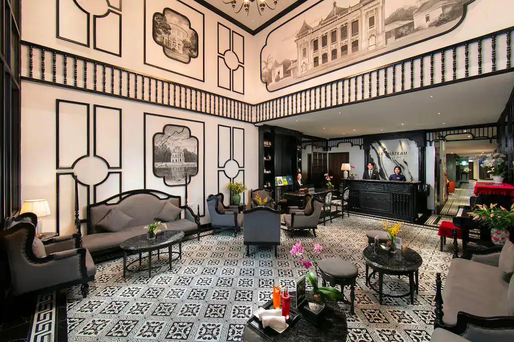 The elegant French colonial lobby of Le Chateau Hotel and Spa in Hanoi, Vietnam, featuring black and white patterned tile floors, ornate wall murals, dark wood furnishings, and a staffed reception desk beneath a sweeping mezzanine balcony.