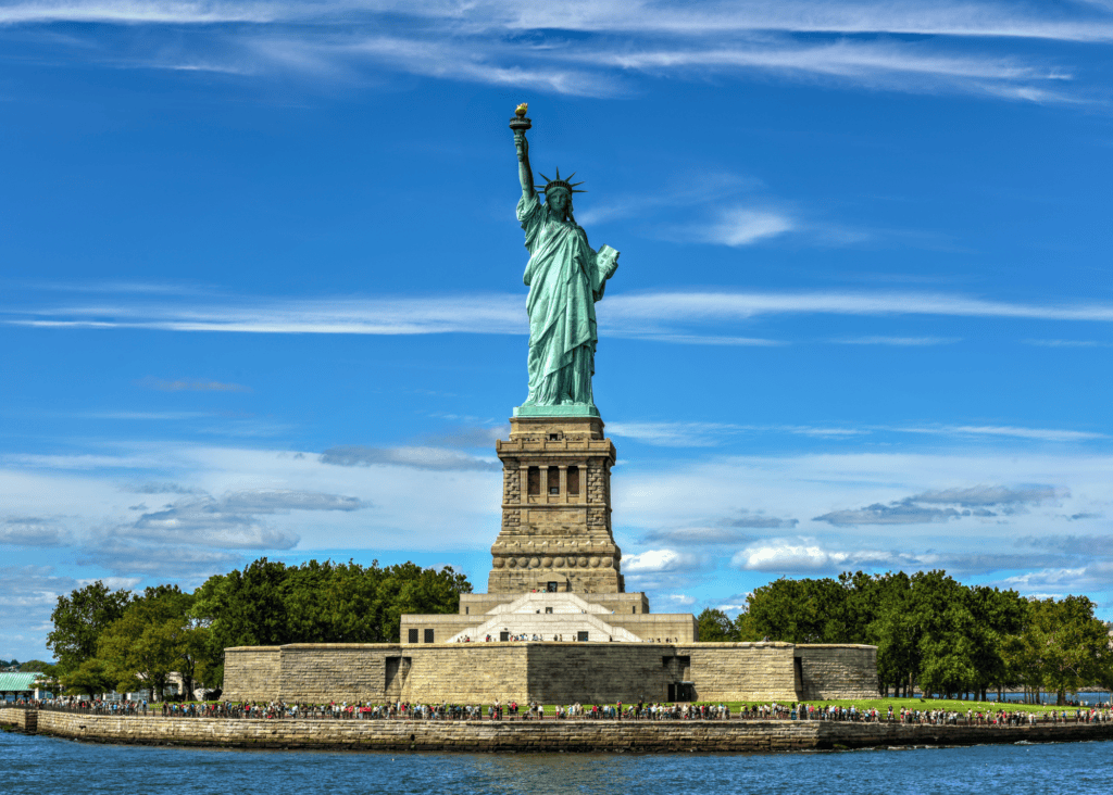 The Statue of Liberty stands tall on Liberty Island in New York Harbor surrounded by crowds of visitors along the base, her torch raised against a bright blue summer sky.