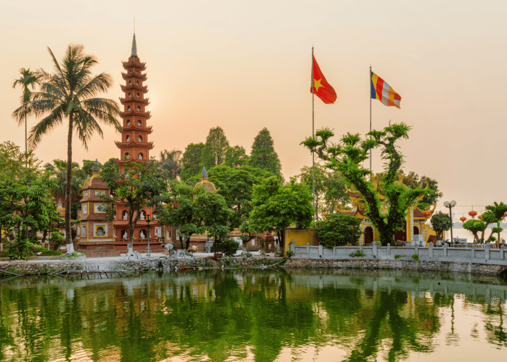 The ancient Tran Quoc Pagoda rises above the still green waters of West Lake in Hanoi, Vietnam, its multi-tiered brick tower glowing warm orange in the soft light of sunset alongside the Vietnamese flag.