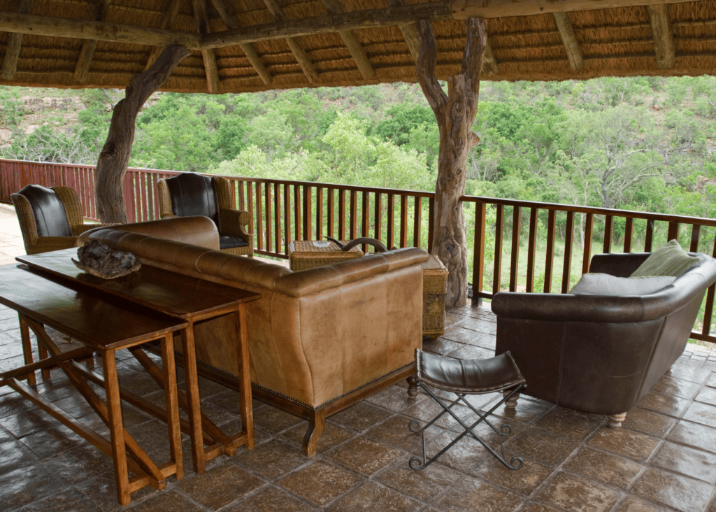 An open air lounge area at a luxury African safari lodge featuring leather sofas, wooden accent tables, and a thatched roof supported by natural tree trunk pillars, with a wooden railing deck overlooking dense green bush beyond.