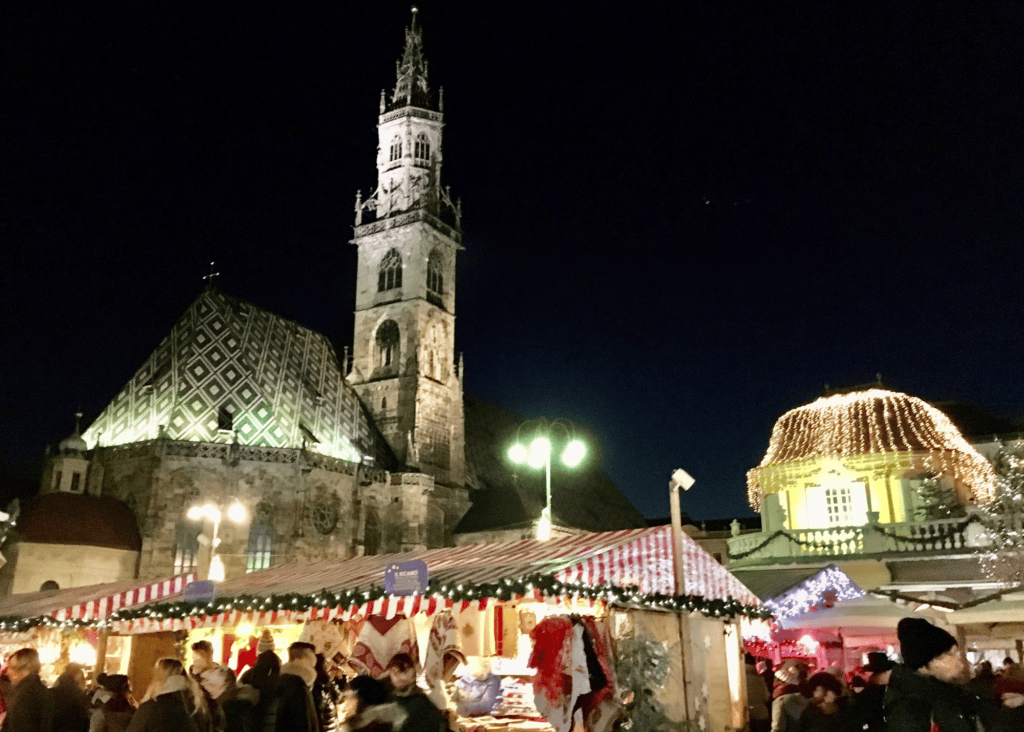 Festive Christmas market stalls with red and white striped rooftops fill the square in front of the illuminated Bolzano Cathedral in South Tyrol, Italy, on a clear winter night.