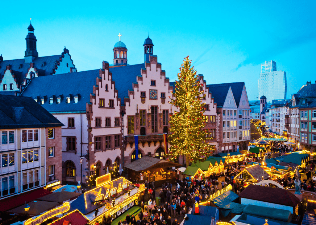 An aerial view of the Frankfurt Christmas market at dusk, with rows of illuminated market stalls and a towering decorated tree filling the historic Römerberg square in front of the medieval city hall.