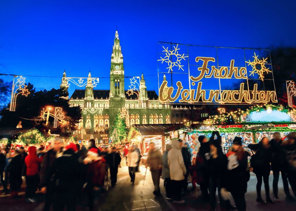Crowds of visitors move through the Vienna Christkindlmarkt at twilight, passing under a glowing Frohe Weihnachten sign with the illuminated Gothic spires of the Rathaus rising in the background.