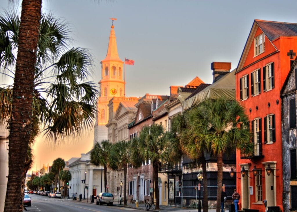 A sunset lit street in historic Charleston, South Carolina, with tall palm trees lining a cobblestone road and a golden hued church steeple rising above rows of colorful antebellum buildings, including a vivid orange facade, as the evening sky glows soft gray and gold.