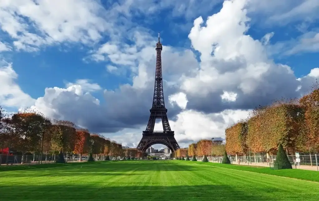 Eiffel Tower rising above the green lawns of Champ de Mars in Paris France on a sunny day with autumn trees and dramatic clouds.
