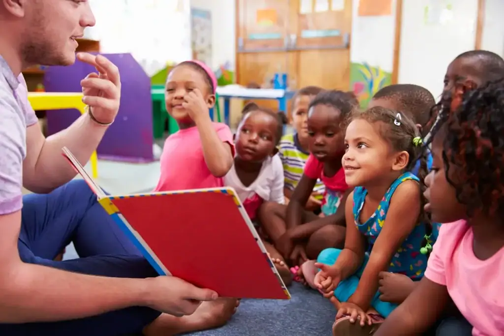 A volunteer reads aloud from a colorful picture book to a group of young children seated on the floor in a bright classroom. The children are engaged and smiling, some leaning in with curiosity.