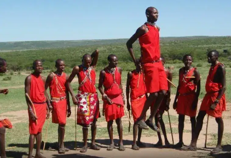 A group of Maasai warriors dressed in vibrant red shukas and colorful beaded jewelry perform a traditional adumu jumping dance outdoors, with one warrior mid-leap above the group against a backdrop of green rolling hills and blue sky in Kenya.