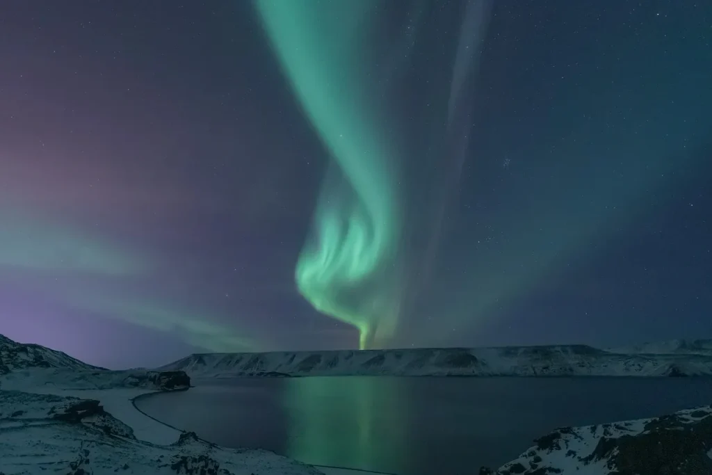 Swirling ribbons of green and teal aurora borealis rise dramatically above a calm arctic fjord, with snow covered cliffs lining the shoreline and the Northern Lights reflecting softly on the still water below.