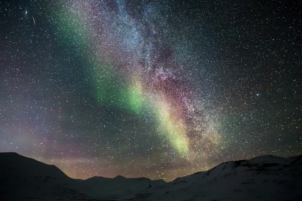 A breathtaking night sky illuminated by the Northern Lights, with sweeping bands of green and violet aurora borealis stretching across a star filled sky above a dark silhouetted mountain horizon.