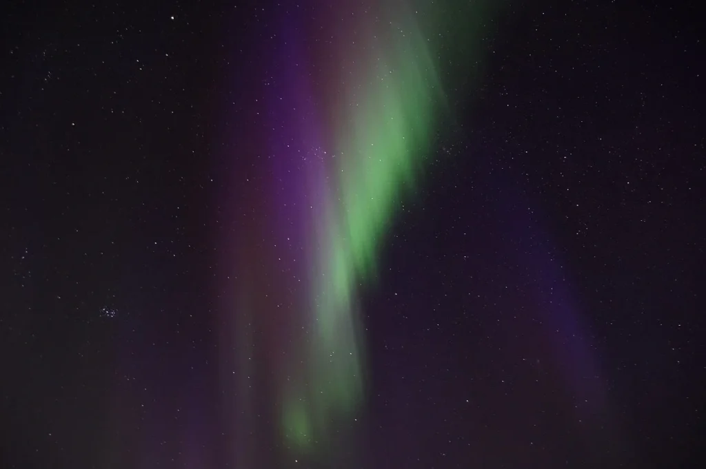 Tall vertical columns of green and purple aurora borealis illuminate a deep black starlit sky, showcasing the vivid color range of a strong Northern Lights display with no horizon visible, just pure sky and light.