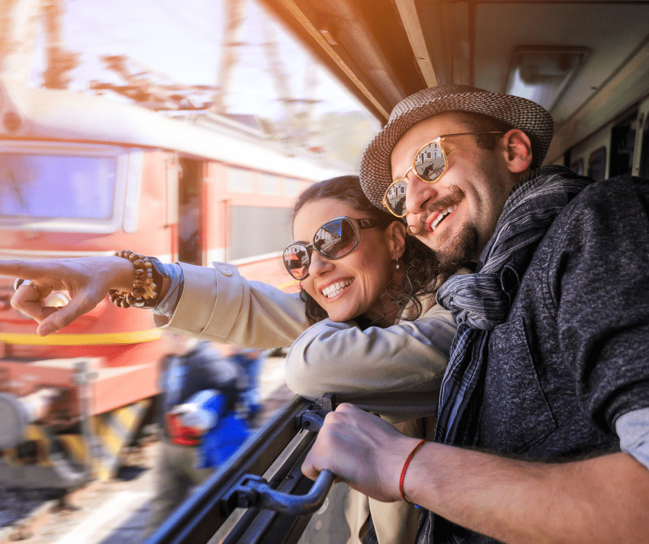 A smiling couple leans out together, one pointing excitedly into the distance, both wearing sunglasses and casual travel attire. The warm, candid moment captures the joy of exploring somewhere new together.
