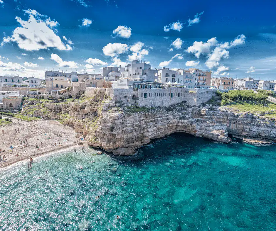 Whitewashed trulli buildings and olive groves in Puglia, Italy, with a clear turquoise sea visible in the distance