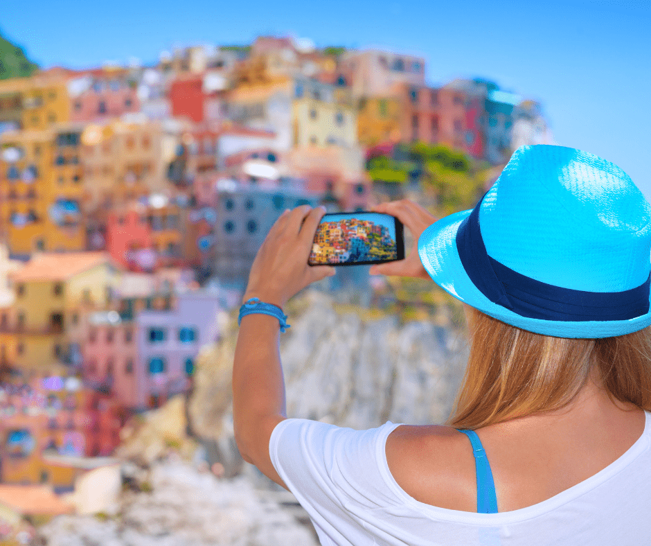 Woman in a blue hat photographing the colorful cliffside village of Manarola in Cinque Terre Italy with her smartphone on a sunny day.