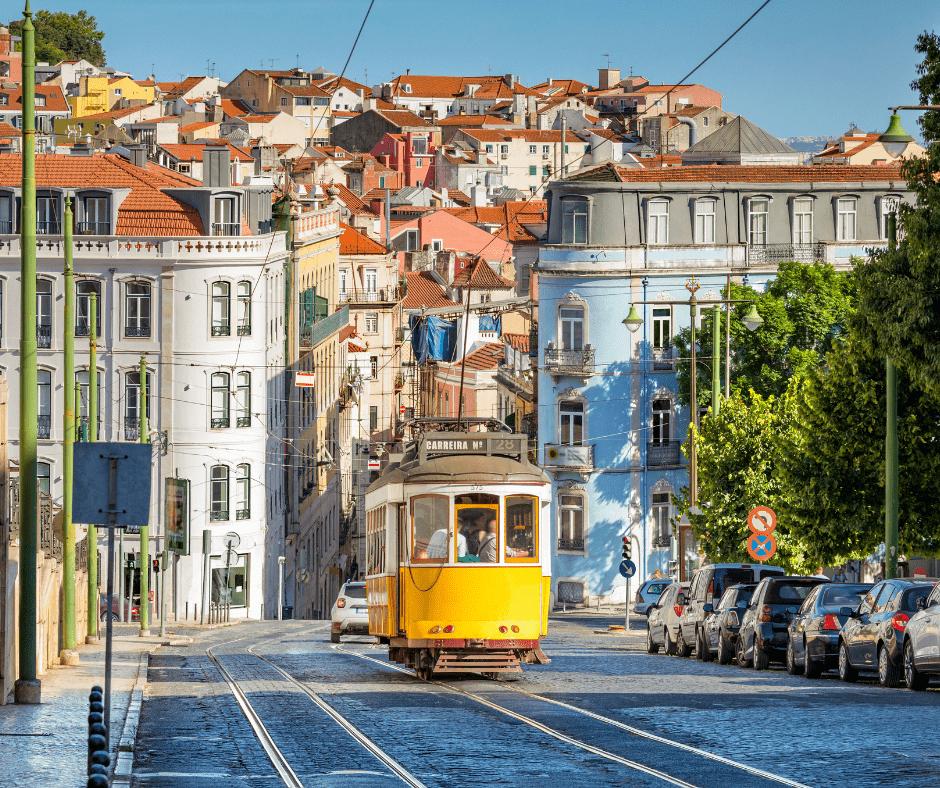 Iconic yellow tram climbing a cobblestone hill in Lisbon Portugal with colorful buildings and red rooftops in the background.