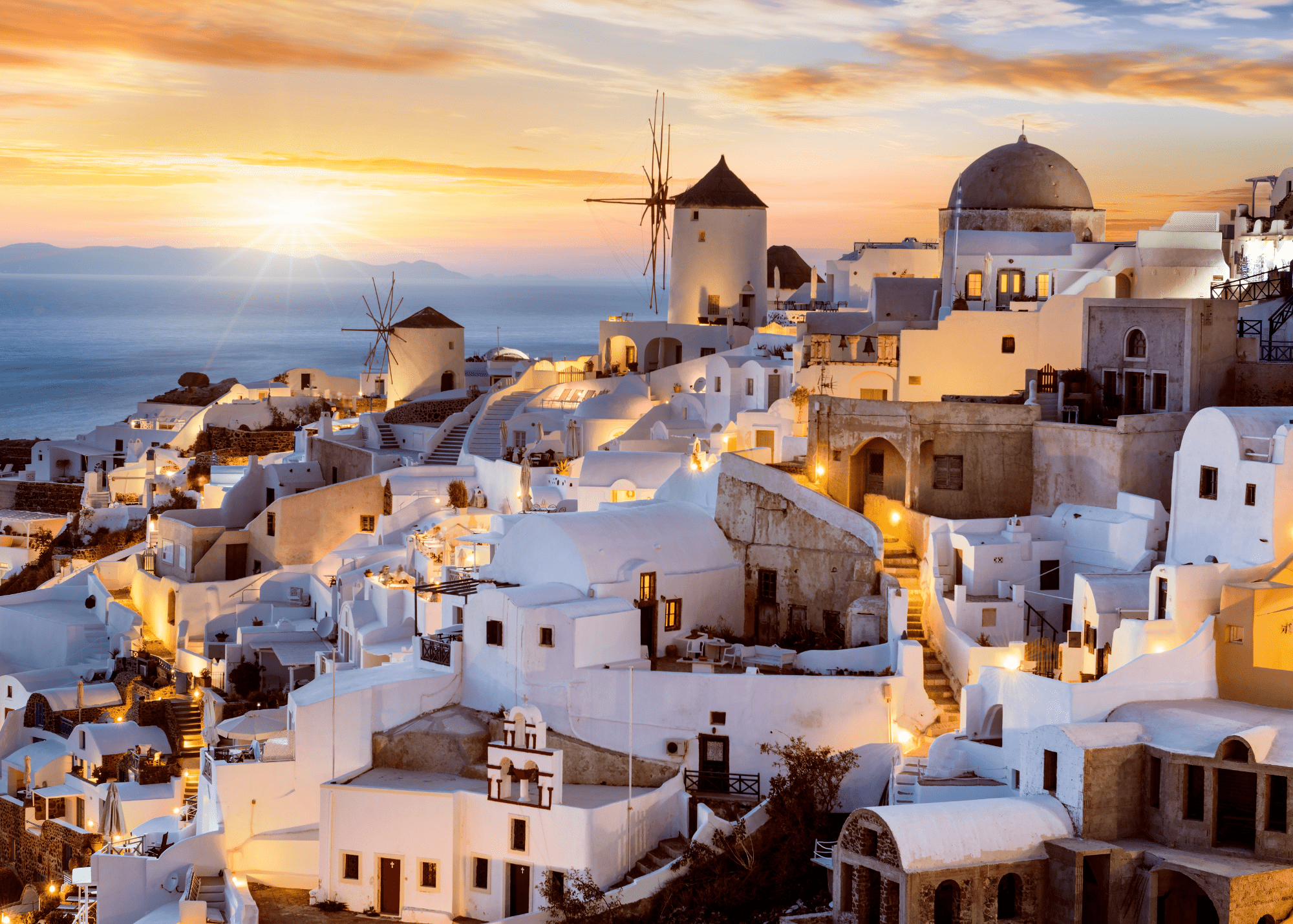 White washed buildings and windmills of Oia village in Santorini Greece glowing in warm golden sunset light overlooking the Aegean Sea.