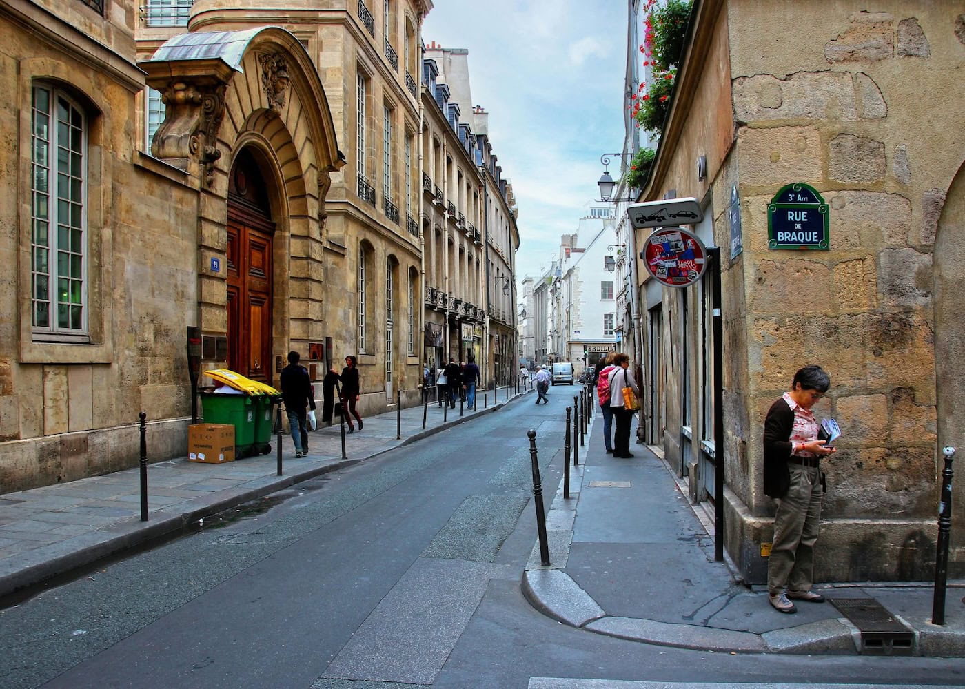 Narrow cobblestone street in the Marais district of Paris France with classic Haussmann stone buildings, pedestrians walking, and a green street sign reading Rue de Braque on the corner