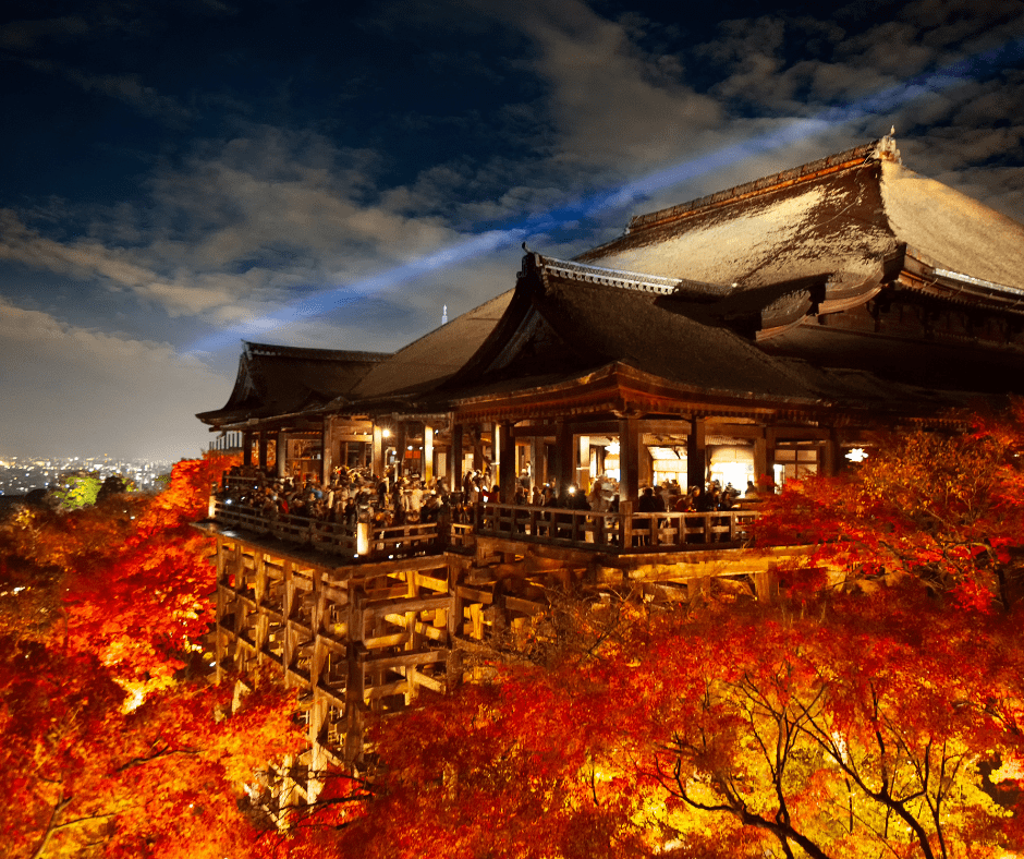 Kiyomizu-dera Temple wooden stage lit up at night surrounded by vibrant red and orange autumn foliage with Kyoto city lights in the background