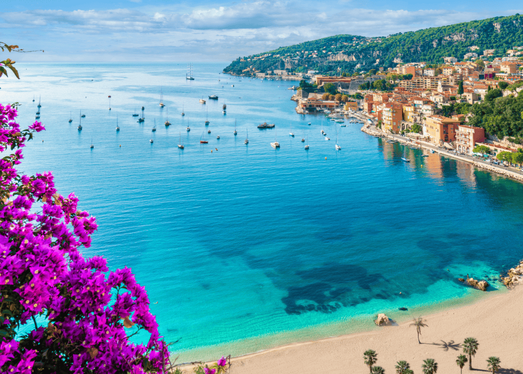 Villefranche-sur-Mer bay on the French Riviera with turquoise water, sailboats, and pink bougainvillea in the foreground