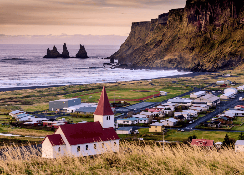 Aerial view of the small coastal village of Vik Iceland with a red roofed church, black sand beach, dramatic sea stacks, and cliffs at sunset.