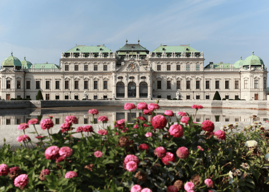 Belvedere Palace in Vienna Austria reflected in a still pool with pink flowers blooming in the foreground on a clear day.