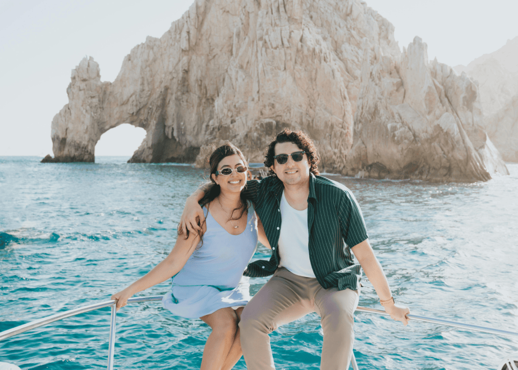 A happy couple sits together on the bow of a boat with the iconic stone arch of Cabo San Lucas rising from the turquoise sea behind them.