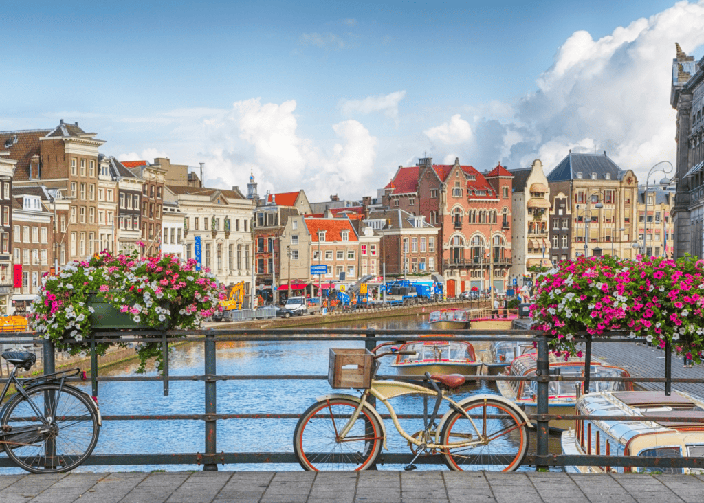 Bicycle and pink flower boxes on a canal bridge in Amsterdam Netherlands with historic row houses and boats in the background.