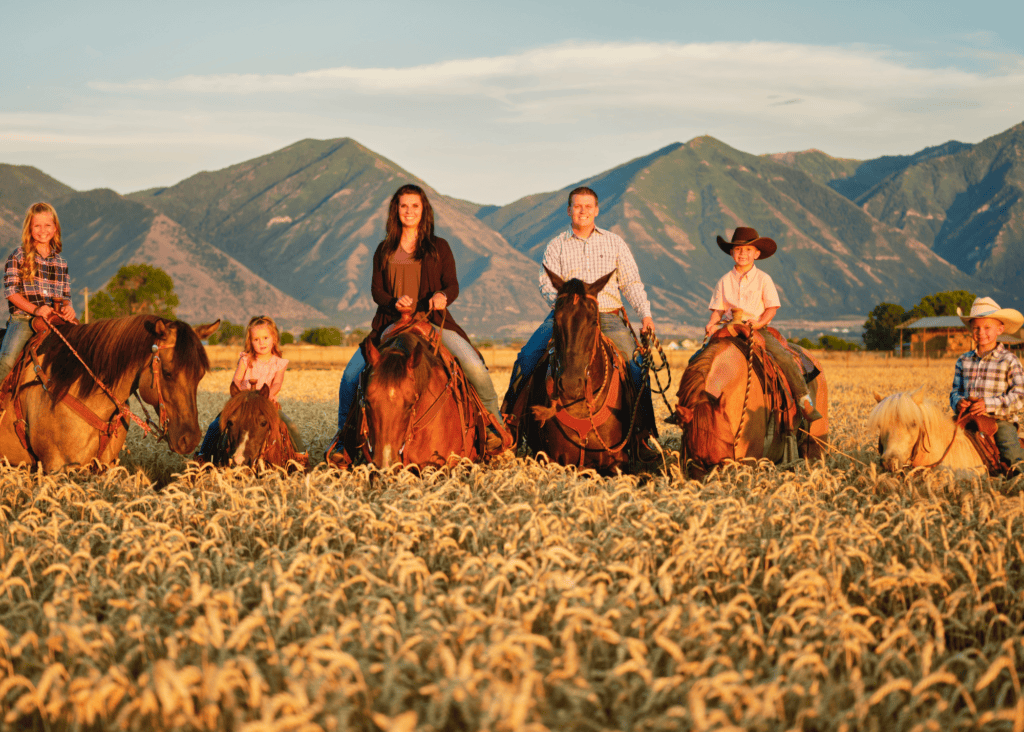 A family of five on horseback riding through a golden wheat field at sunset with mountain ranges in the background at a dude ranch in the American West.
