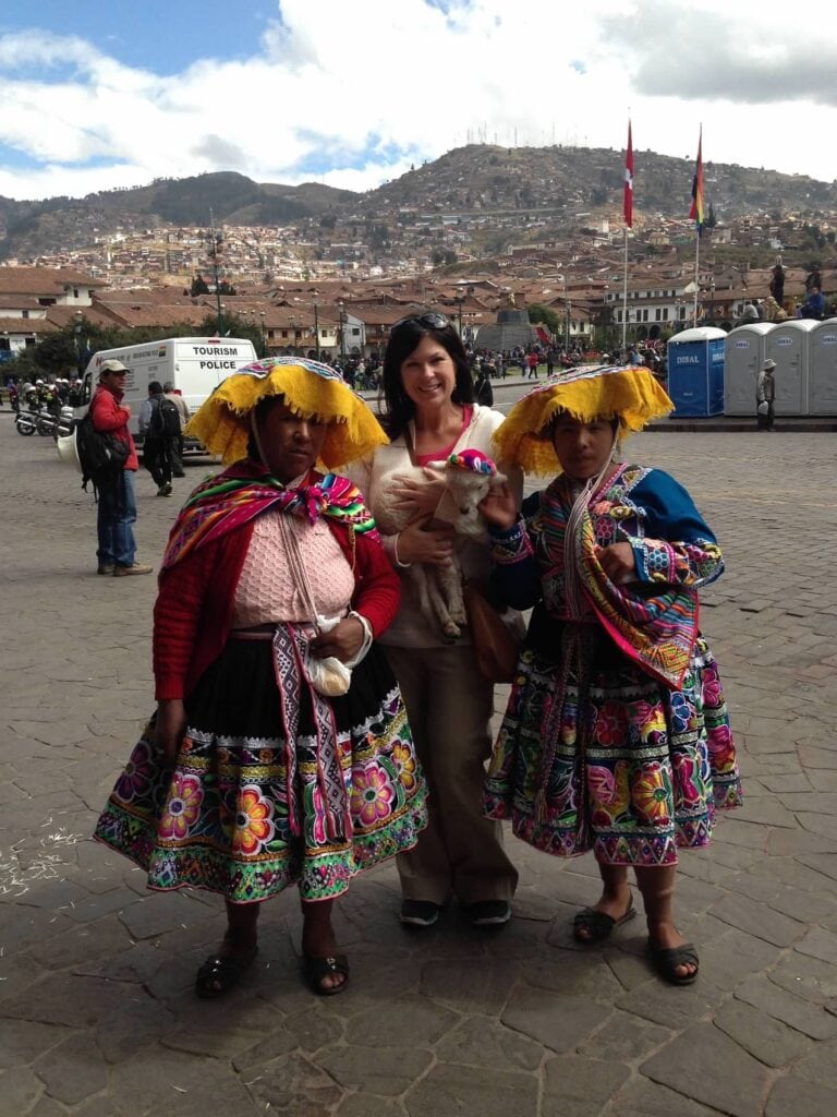 Gina Arefi standing in the main plaza of Cusco Peru holding a baby sheep flanked by two women in vibrant traditional Peruvian dress with the city in the background.