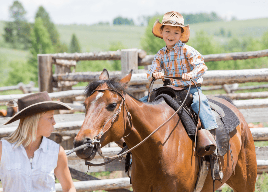 A young boy in a cowboy hat smiling while riding a chestnut horse at a dude ranch, guided by a wrangler at the corral fence.