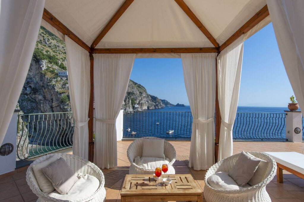 Shaded white canvas cabana on the terrace at Onda Verde in Praiano Italy with white wicker chairs, two Aperol Spritzes on a wooden table, and dramatic views of the Amalfi Coast cliffs and the Tyrrhenian Sea through the open curtains.