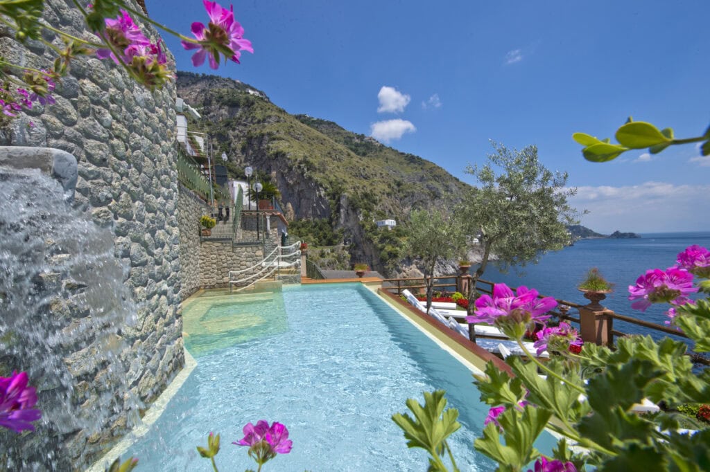 Infinity pool at Onda Verde in Praiano Italy with a stone waterfall feature, pink geraniums in the foreground, olive trees, and sweeping views of the Amalfi Coast cliffs and Tyrrhenian Sea.