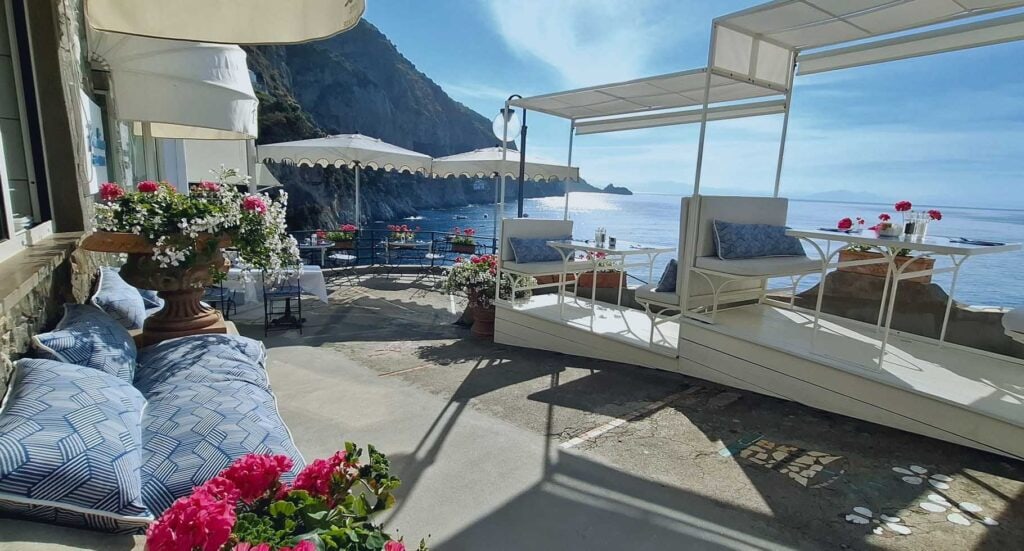 Daytime view of the restaurant terrace at Onda Verde in Praiano Italy with white dining cabanas, blue cushions, pink and white geraniums in terracotta urns, and the Amalfi Coast cliffs and sparkling Tyrrhenian Sea in the background.