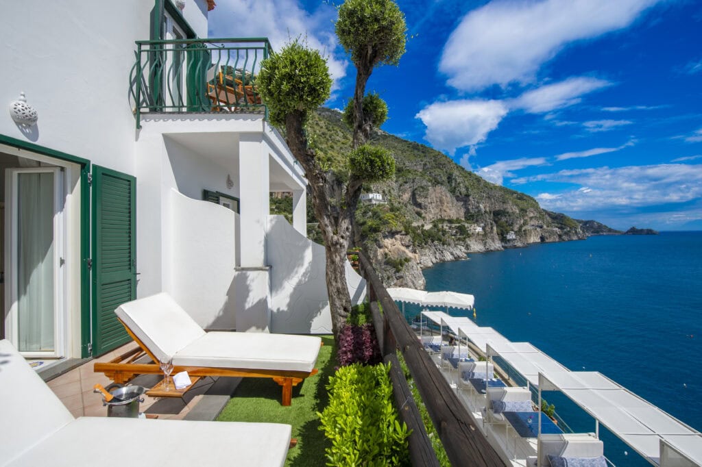 Private terrace at Onda Verde in Praiano Italy with teak loungers, a champagne bucket, and sweeping views of the dramatic Amalfi Coast cliffs and the deep blue Tyrrhenian Sea with the restaurant terrace visible below.