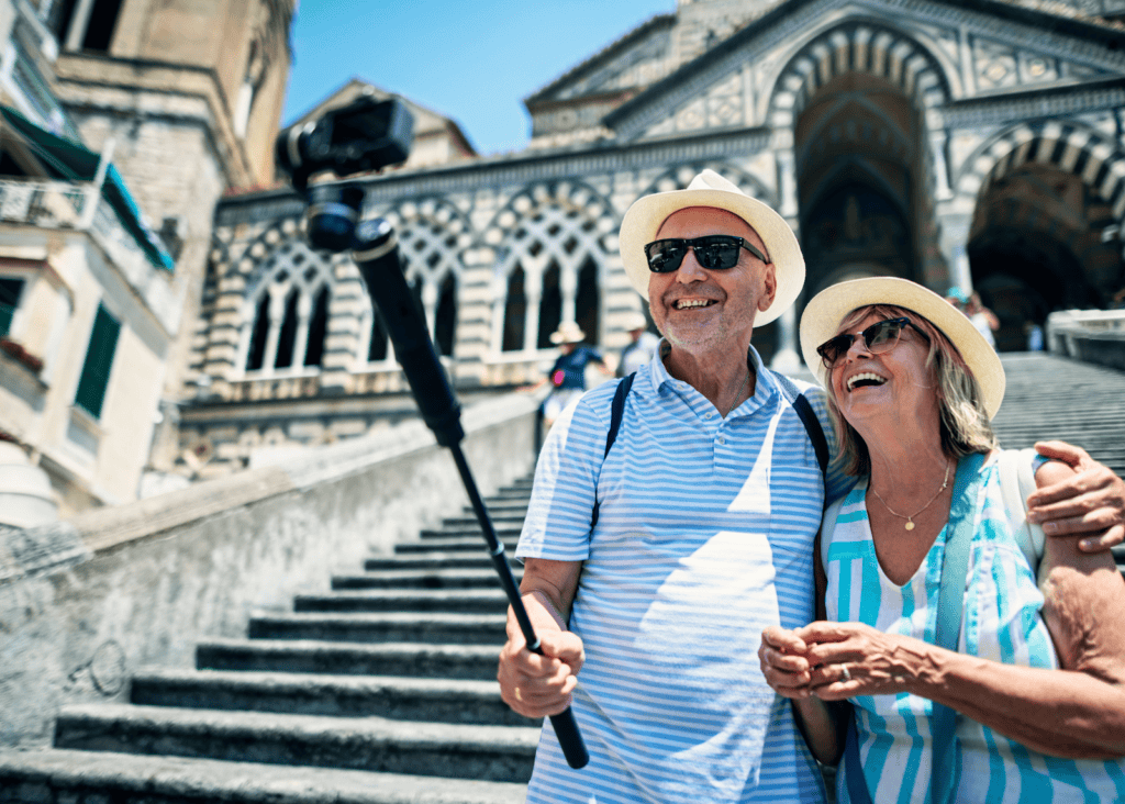 A joyful older couple takes a selfie on a selfie stick in front of the ornate facade of the Amalfi Cathedral on a bright sunny day.