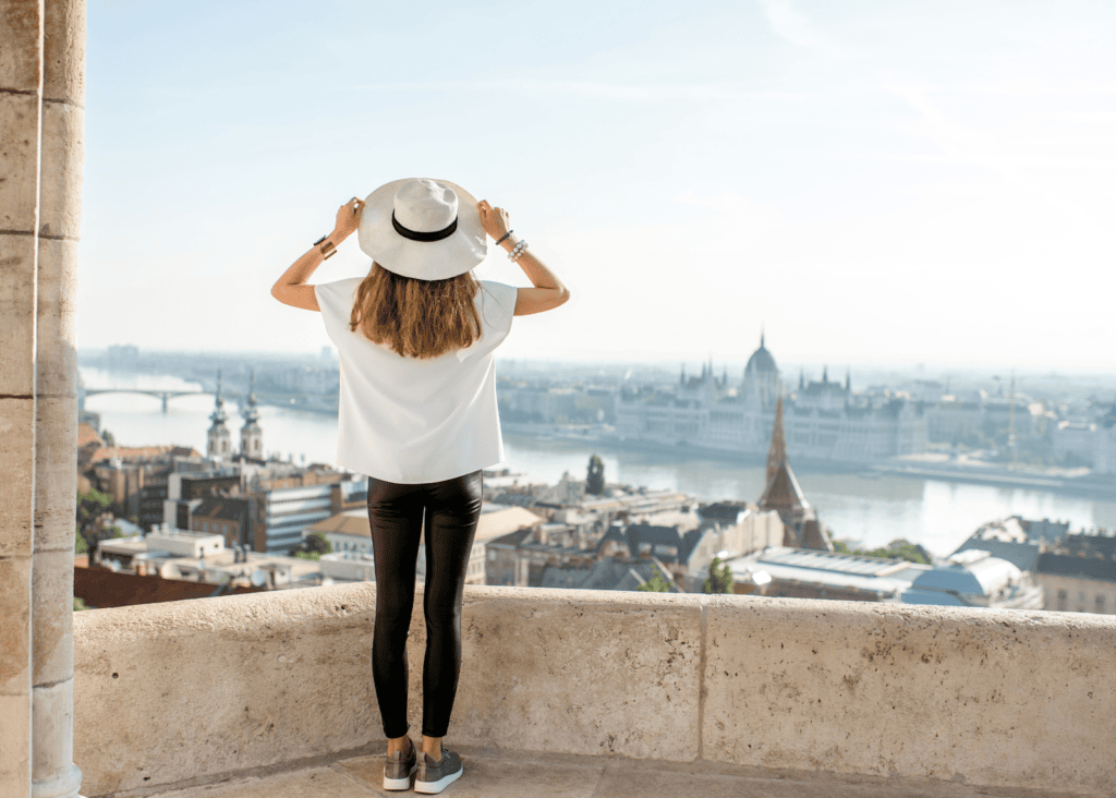 A solo female traveler stands with her back to the camera at a stone overlook, holding her white hat and gazing out over the Budapest skyline and the Danube River below.