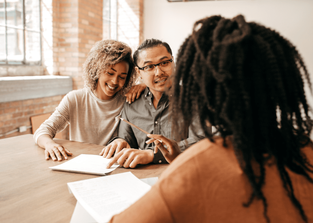 A smiling couple sits across from a travel advisor at a wooden table, reviewing trip documents together in a warm, brick-walled setting.