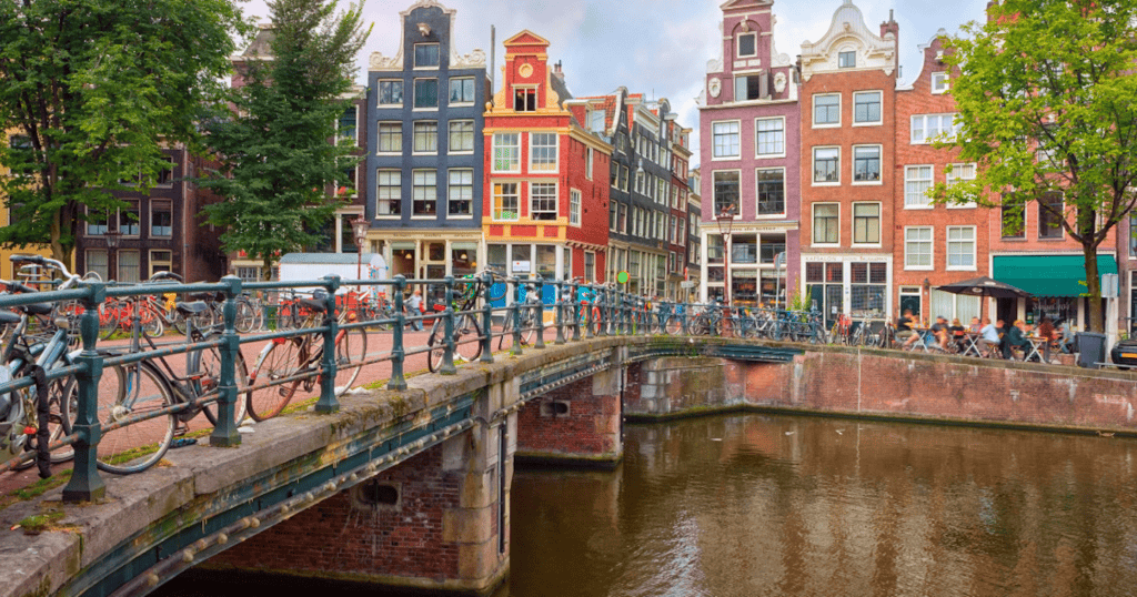 Bicycles line a brick bridge over an Amsterdam canal with colorful narrow canal houses and a waterfront cafe visible in the background on a cloudy summer day.