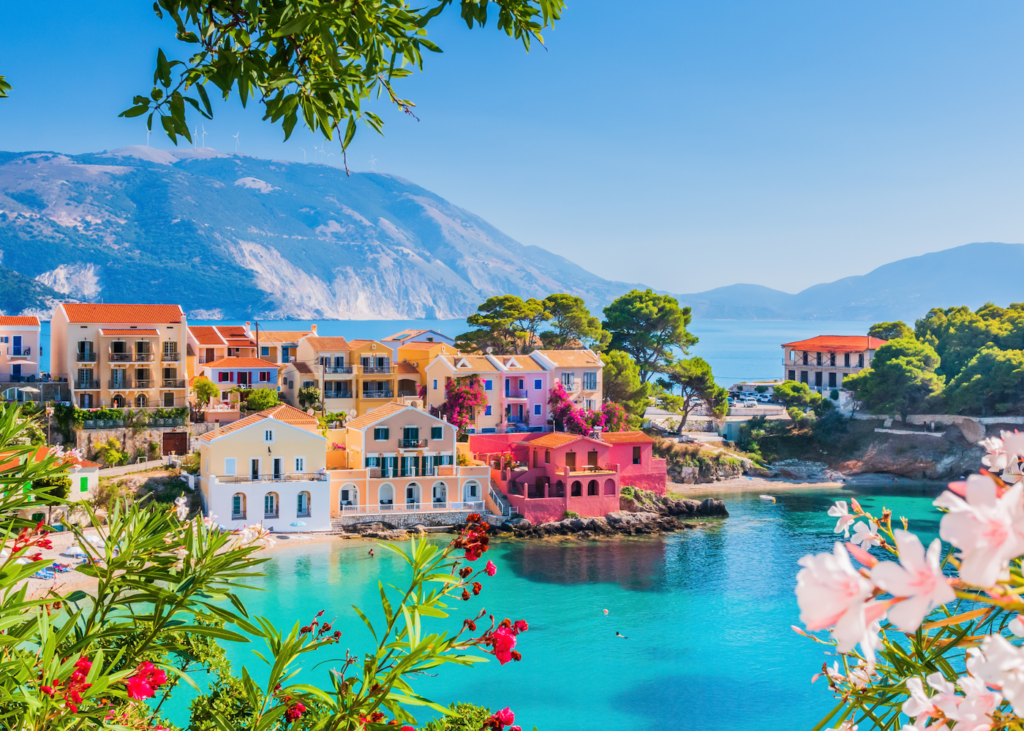 The colorful village of Assos in Kefalonia Greece sits along a turquoise bay framed by pink and red flowering oleander with mountains rising behind the pastel buildings.