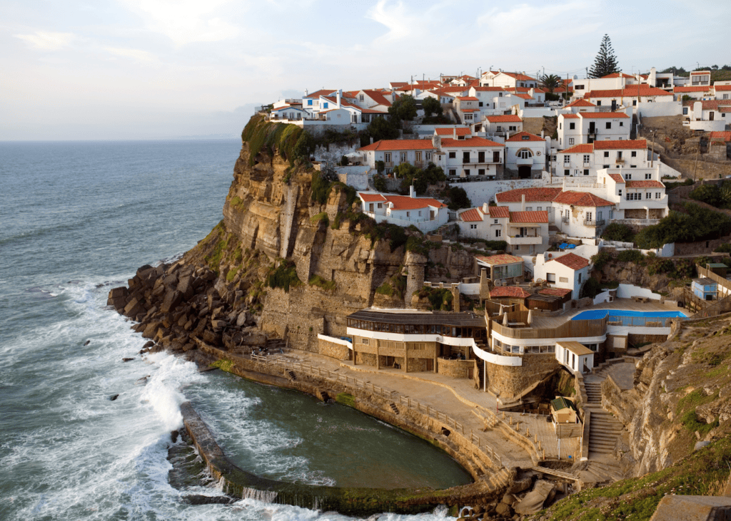 The cliffside village of Azenhas do Mar Portugal with white and terracotta roofed houses perched dramatically above the Atlantic Ocean with crashing waves below and a natural ocean pool carved into the rocks.